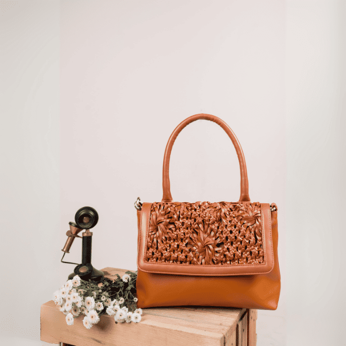 Brown leather vegan leather handbag for women with intricate design on a wooden surface with flowers and a vintage phone.