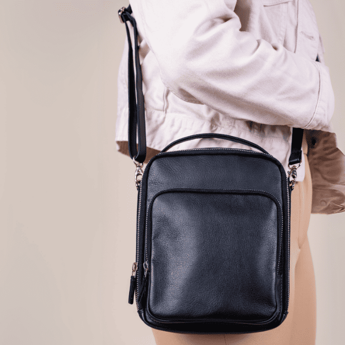Black crossbody vegan leather bag held by a person wearing a light-colored jacket on a beige background