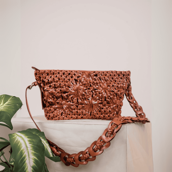 Brown woven leather handbag for women with chain detail on a white surface with a plant in the foreground.