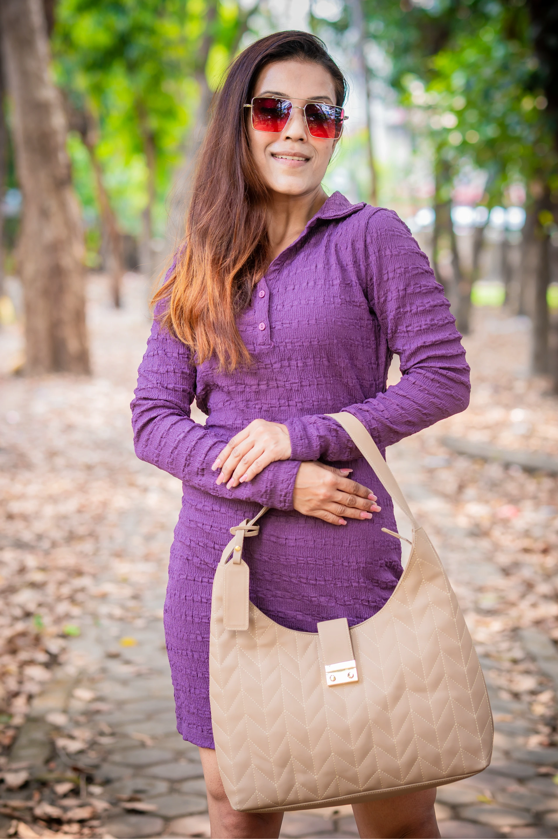 Woman in purple dress holding a beige bag outdoors with greenery in the background