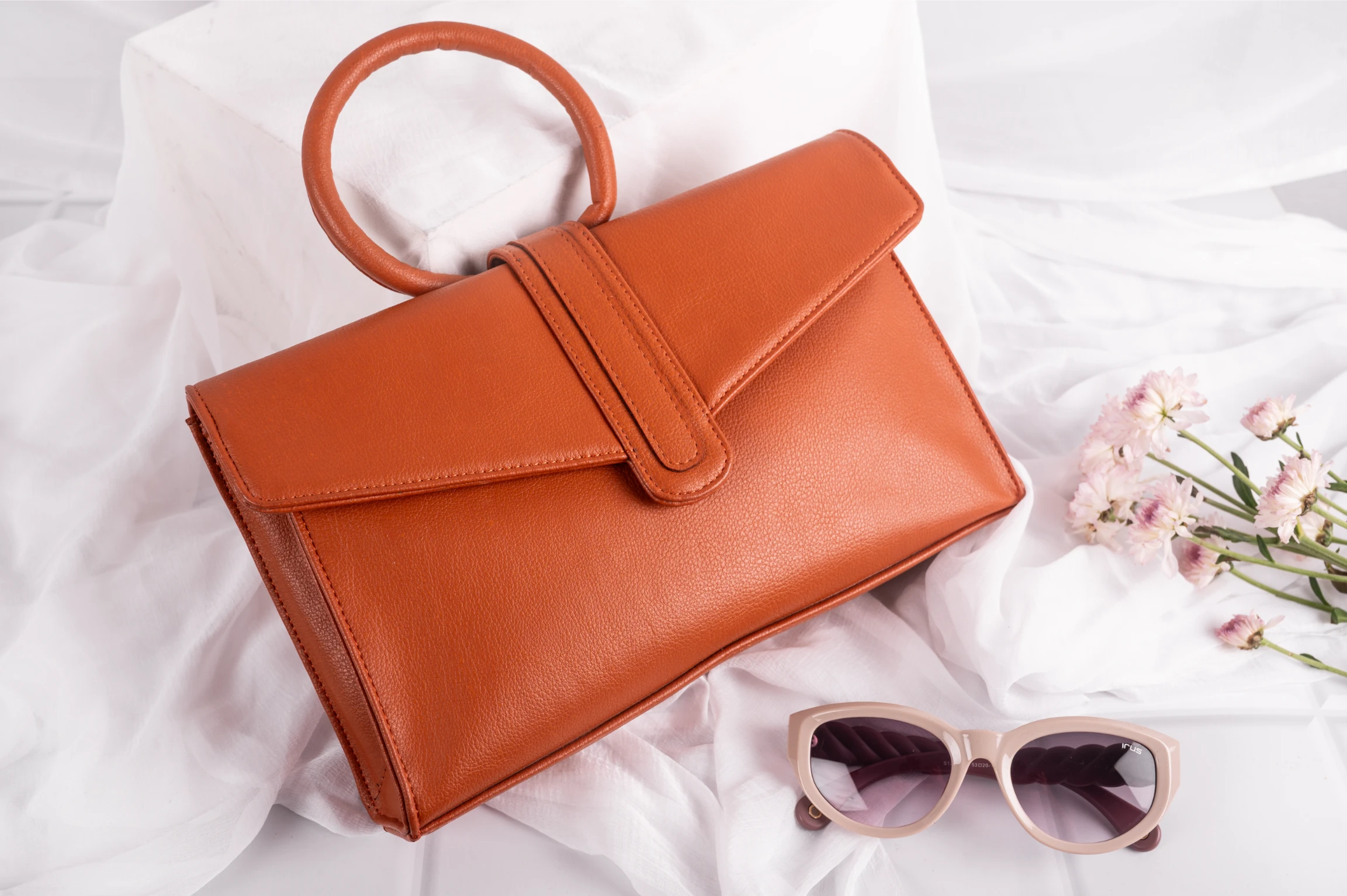 Brown vegan leather handbag for women and sunglasses on a white fabric background with pink flowers.