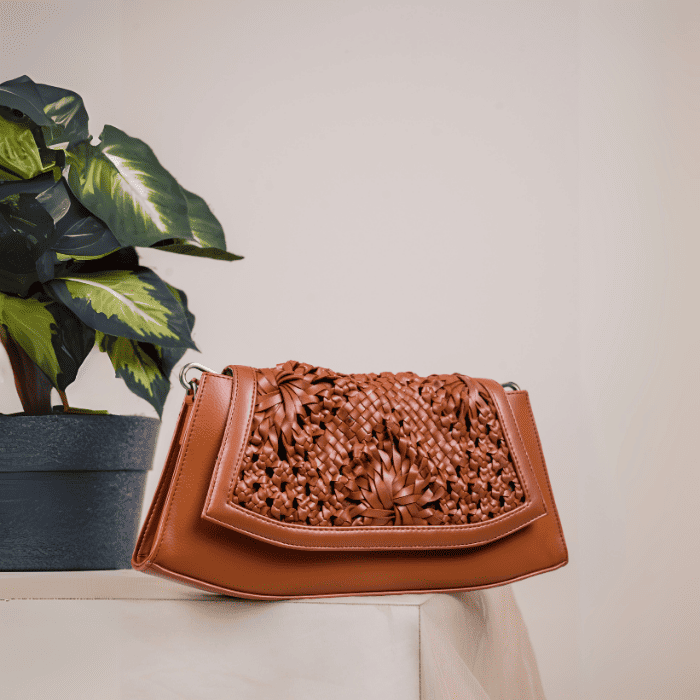 Brown vegan leather handbag for women with intricate design on a white surface next to a plant