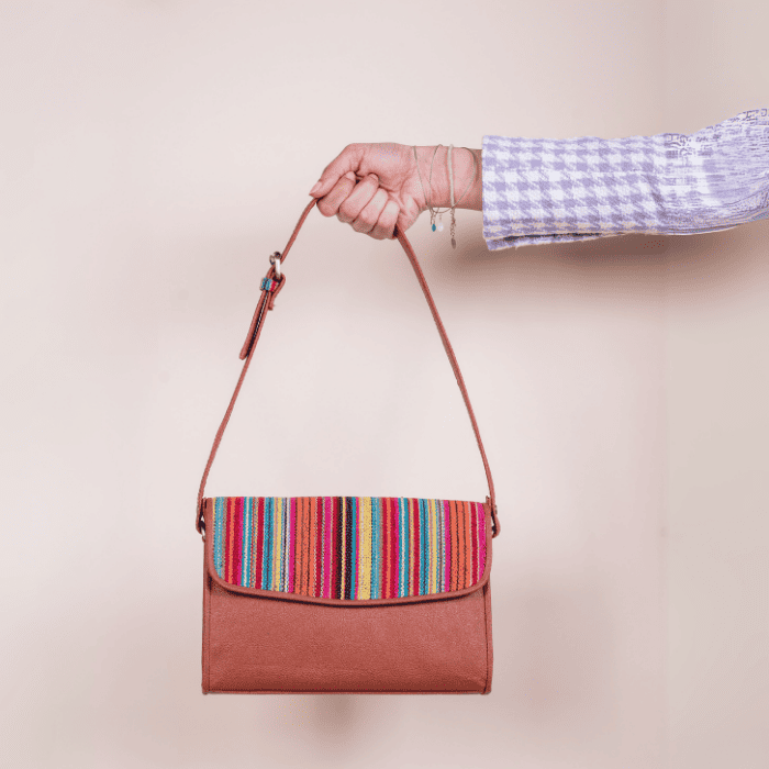 Hand holding a brown vegan leather bag for women with a colorful striped pattern on a beige background