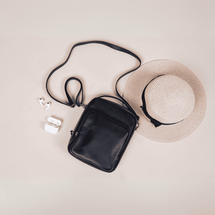 Black vegan leather crossbody bag with a straw hat and white earbuds on a beige background