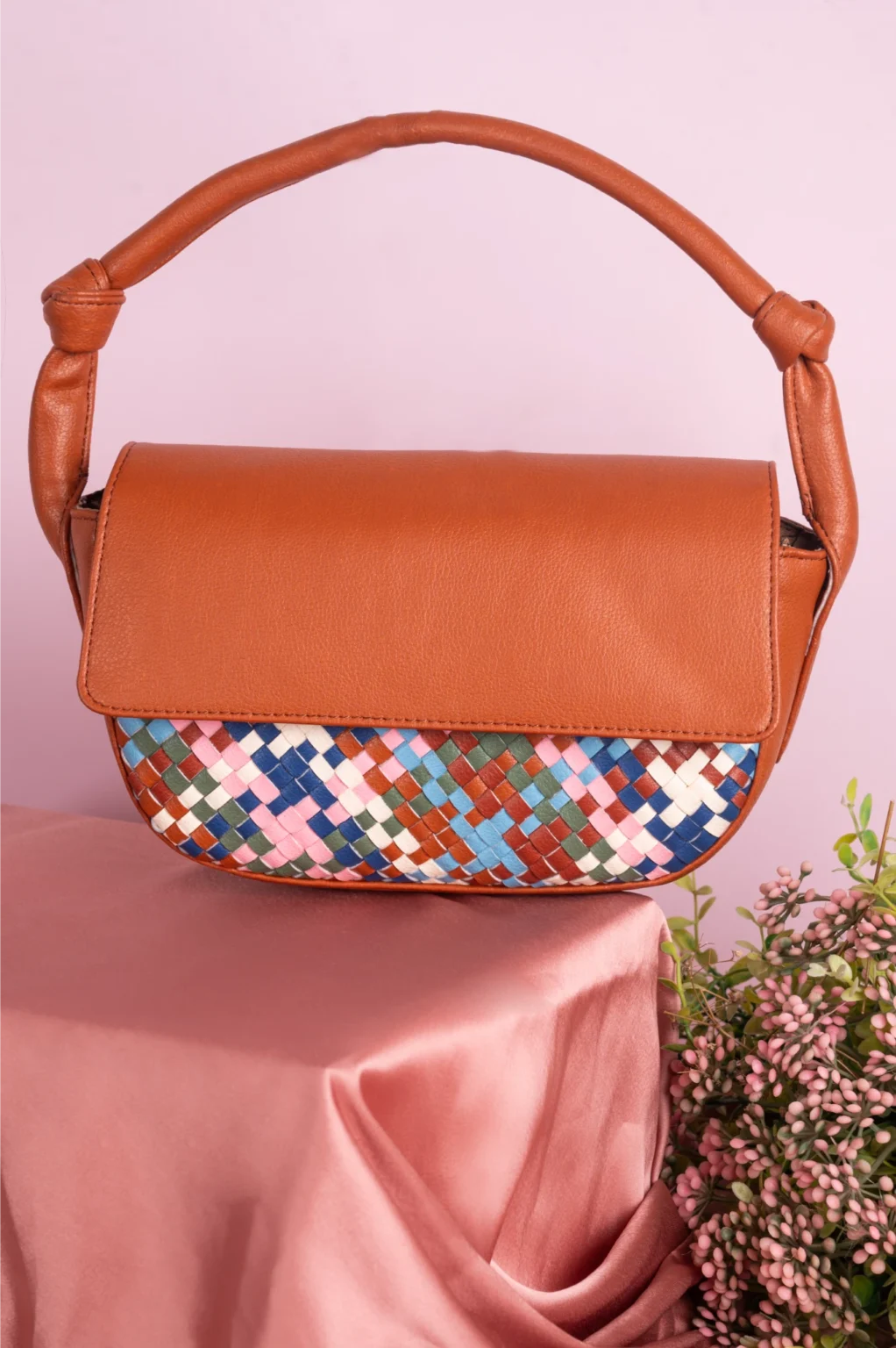 Brown vegan leather handbag for women with a colorful woven pattern on a pink surface with flowers.