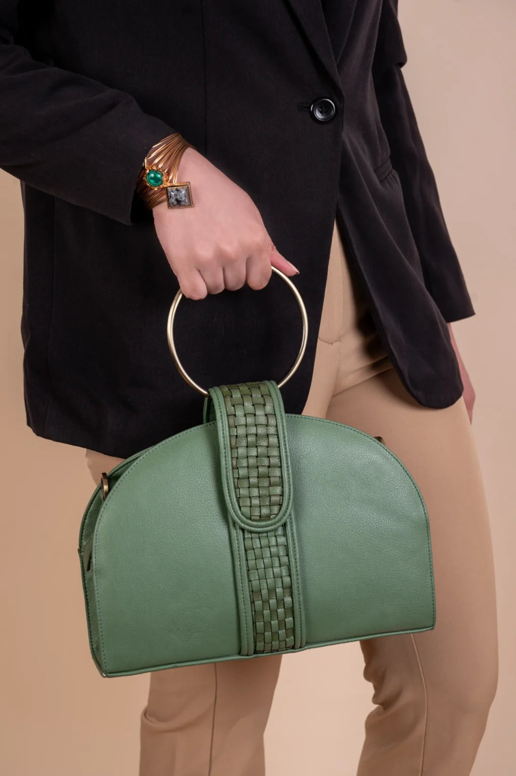 Green vegan leather handbag for women with a circular handle held by a person wearing a black coat and beige pants on a beige background.