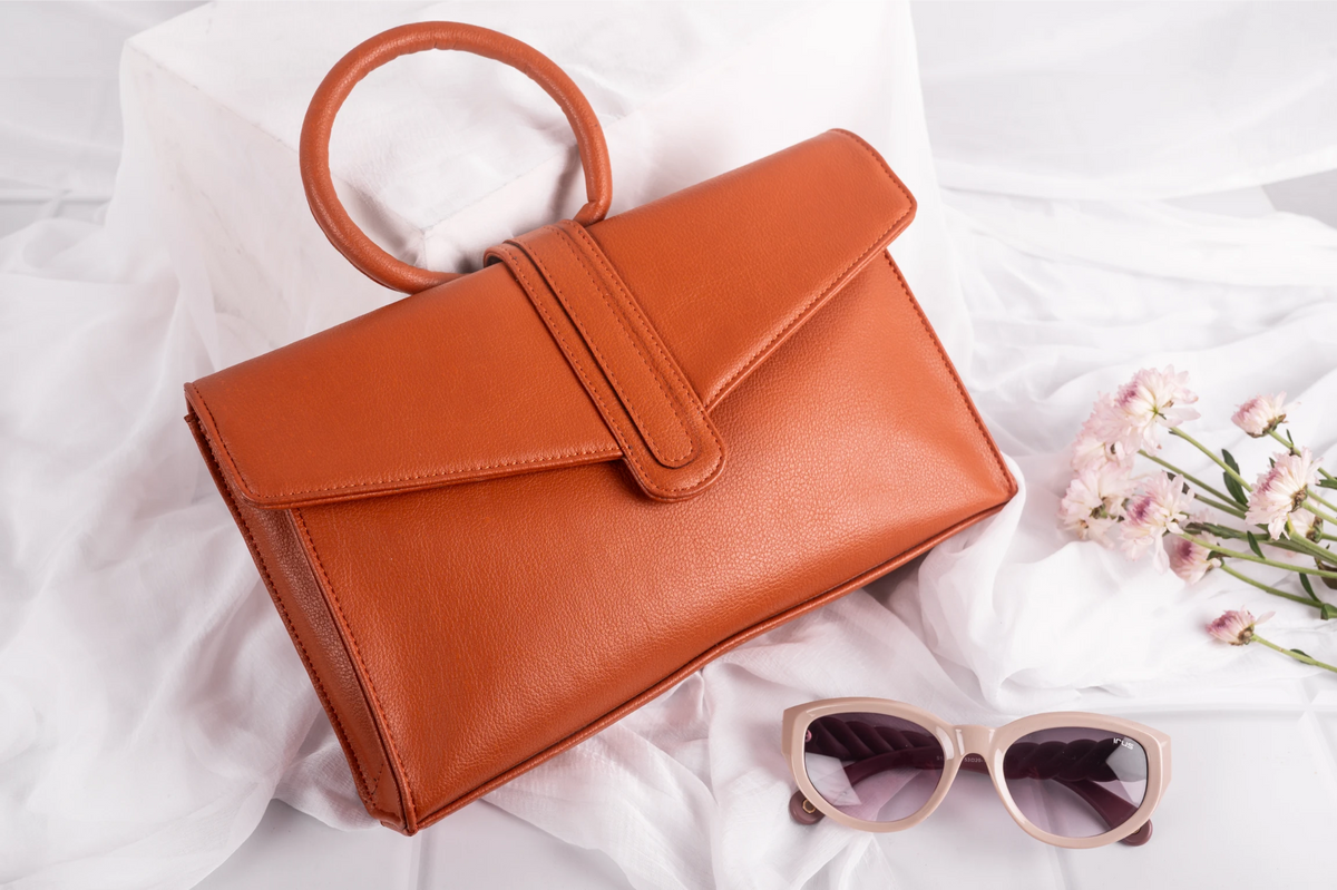 Brown vegan leather handbag for women and sunglasses on a white fabric background with pink flowers.