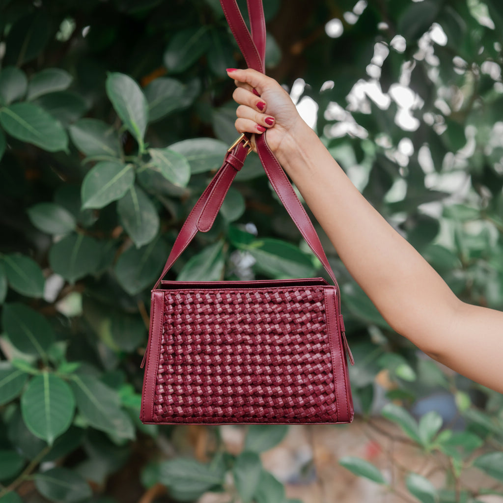 Hand holding a maroon woven vegan leather handbag for women against a blurred green leafy background