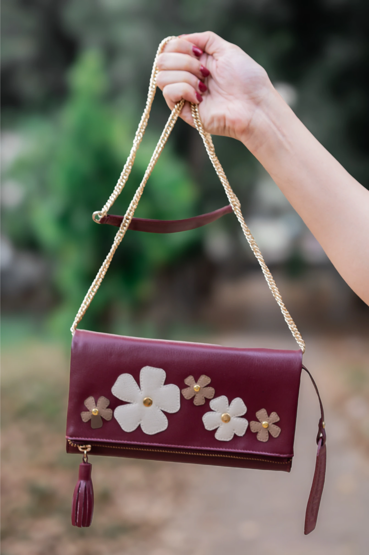 Maroon vegan leather handbag for women with floral designs and a tassel held by a hand against a blurred natural background
