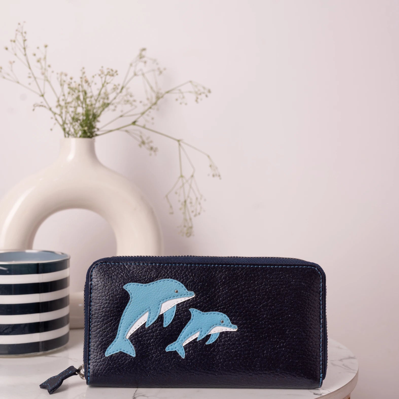 Black vegan leather wallet for women with blue dolphin designs on a white surface with a vase and striped container in the background.