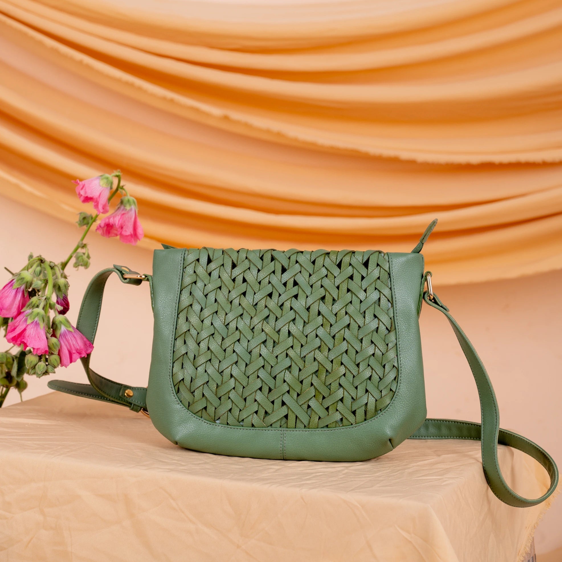 Green woven leather handbag for women on a beige surface with pink flowers and draped fabric in the background