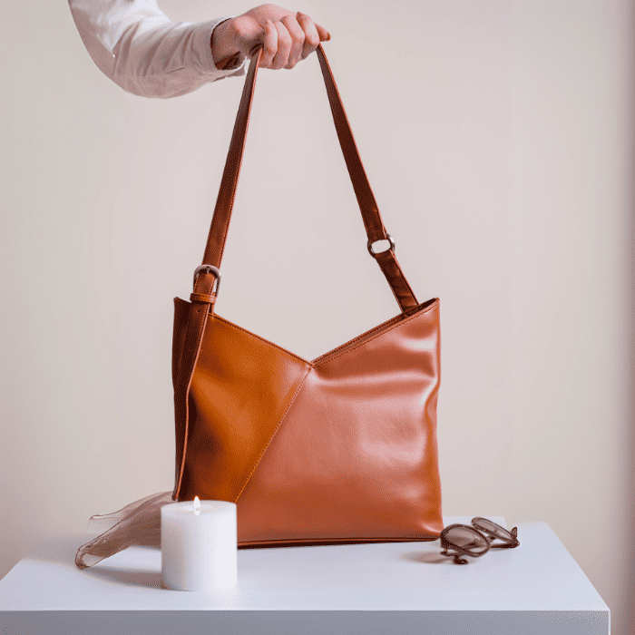 Brown vegan leather handbag for women held by a person on a white surface with a candle and sunglasses.